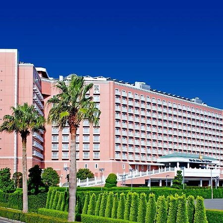 Grand Nikko Tokyo Bay Maihama Hotel Urayasu Exterior foto The image shows a large, multi-story hotel building featuring a light pink facade. The architecture appears modern, with numerous windows arranged in a grid pattern. In the foreground, there are neatly trimmed shrubs and palm trees, adding a tropical