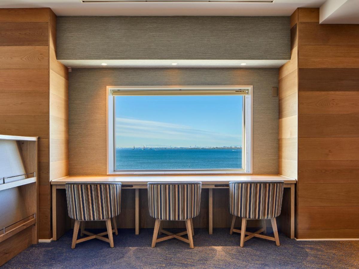 Grand Nikko Tokyo Bay Maihama Hotel Urayasu Exterior foto The photo shows a modern workspace with three striped chairs positioned at a long, white desk. The desk faces a large window with a view of a tranquil body of water and a distant skyline. The walls around the workspace are made of wood paneling, crea