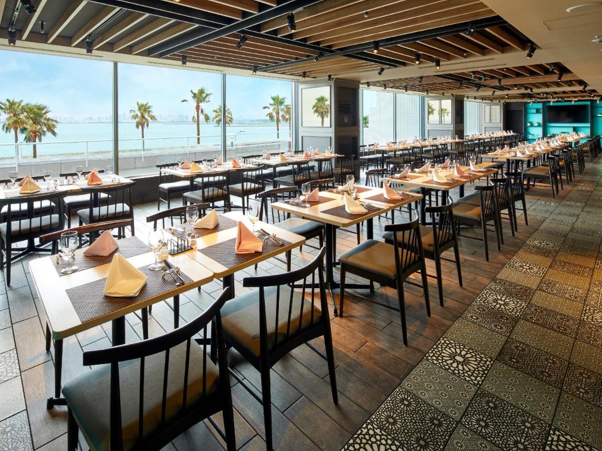 Grand Nikko Tokyo Bay Maihama Hotel Urayasu Exterior foto The photo shows a modern restaurant interior featuring rows of neatly arranged tables and chairs. The large windows provide a scenic view of palm trees and what appears to be a body of water in the background, suggesting a coastal location. Each tabl