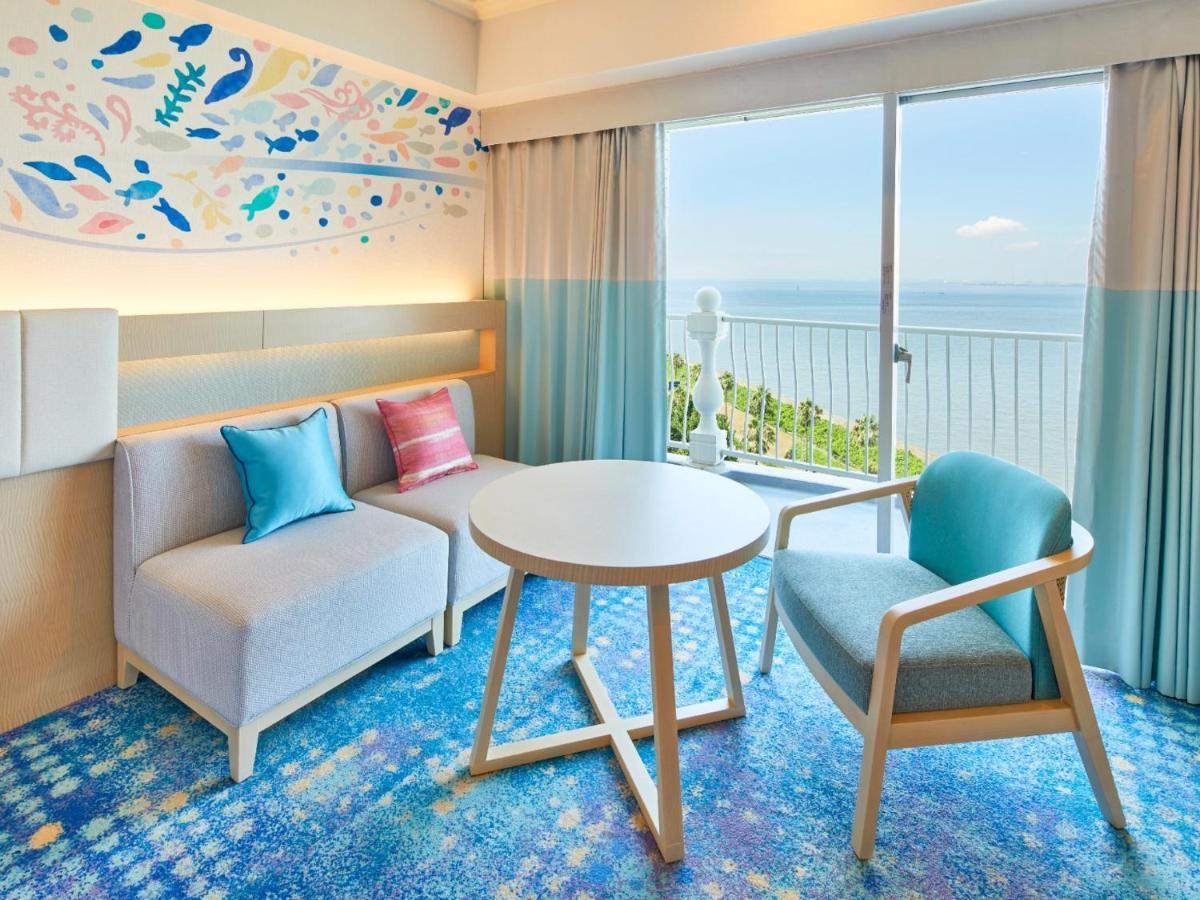 Grand Nikko Tokyo Bay Maihama Hotel Urayasu Exterior foto The photo shows a modern and stylish interior of a room with a view. There is a cozy seating area featuring a light-colored sofa with decorative cushions in various shades of blue and pink. A round table occupies the center of the space, surrounded b