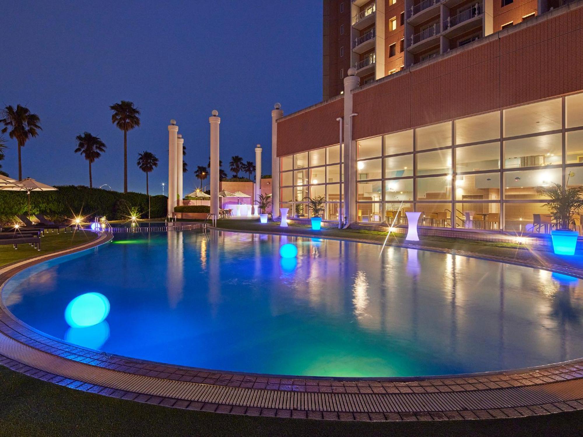 Grand Nikko Tokyo Bay Maihama Hotel Urayasu Exterior foto The photo shows a well-lit swimming pool at night, surrounded by a lush green area. The pool features glowing blue lights that create a serene and inviting atmosphere. Palm trees can be seen in the background, adding a tropical feel to the scene. Add
