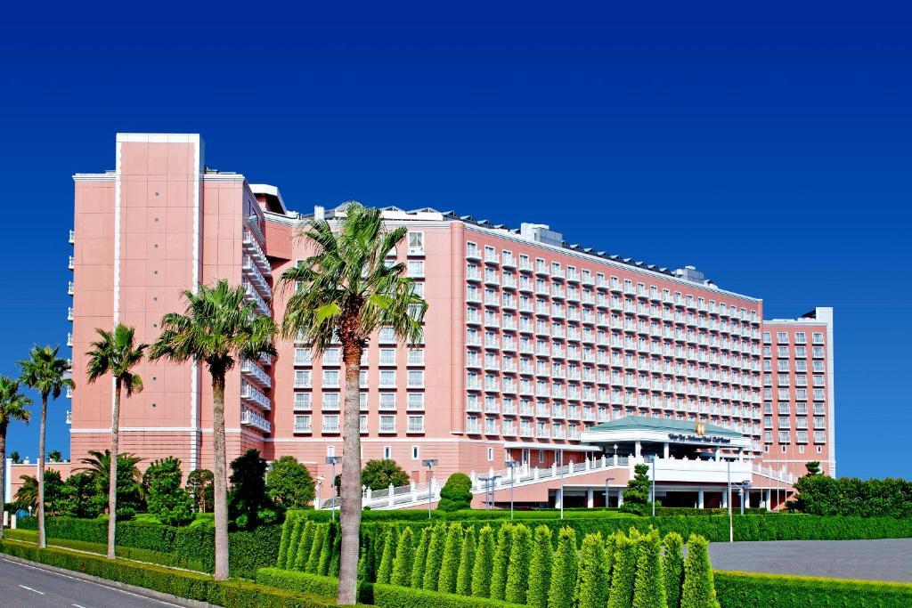 Grand Nikko Tokyo Bay Maihama Hotel Urayasu Exterior foto The image shows a large, multi-story hotel building featuring a light pink facade. The architecture appears modern, with numerous windows arranged in a grid pattern. In the foreground, there are neatly trimmed shrubs and palm trees, adding a tropical