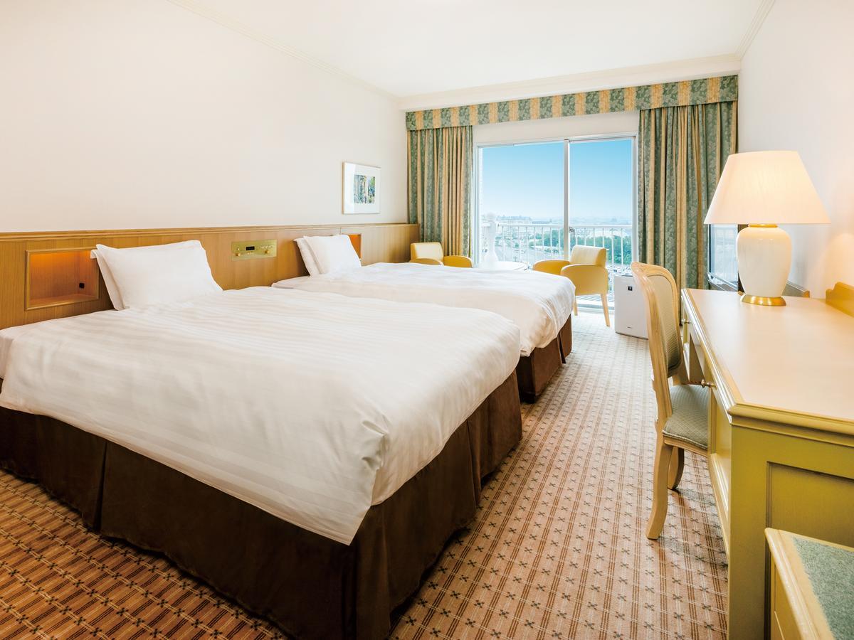 Grand Nikko Tokyo Bay Maihama Hotel Urayasu Exterior foto The photo shows a hotel room featuring two twin beds with white linens and dark brown bed skirts. The room has a light, neutral-colored wall and carpet. There is a window with curtains that let in natural light, offering a view outside. A small desk 